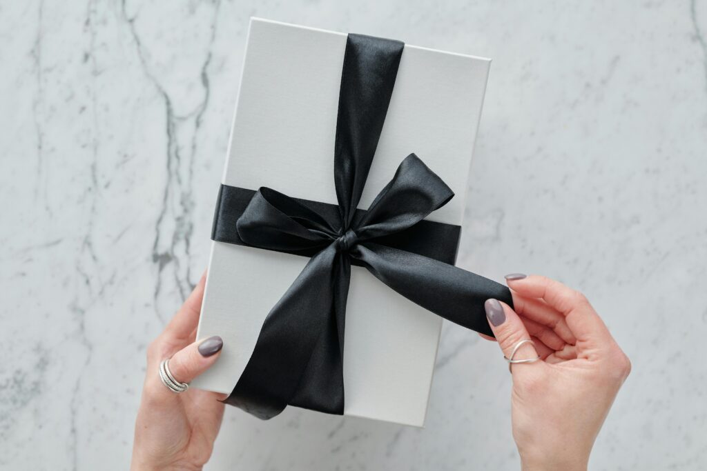 Close das mãos de uma pessoa abrindo uma caixa de presente branca com laço de cetim preto, simbolizando a entrega de pedidos e a experiência de unboxing